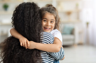 Cute middle-eastern little girl hugging her mother and cheerfully smiling at camera, closeup portrait, panorama with copy space. Happy brunette mom and kid having fun together at home